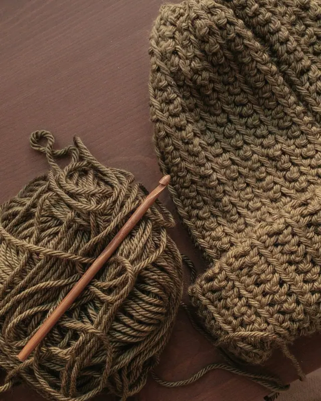 Brown yarn and a wooden crochet hook beside a thick olive crochet piece