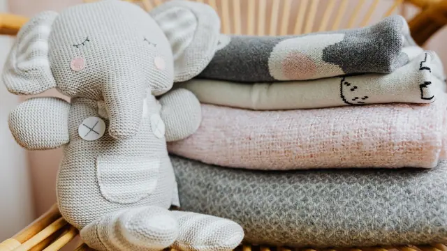 A pale knitted elephant toy beside folded blankets on a wicker chair