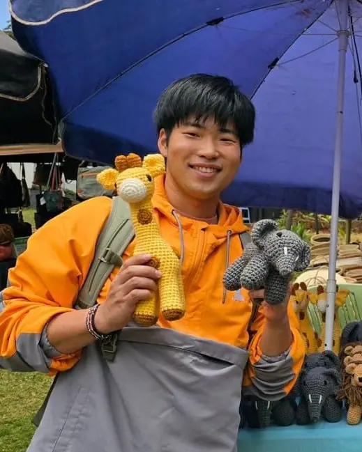 Smiling young man in an orange jacket holding a crocheted giraffe and a grey elephant under a blue umbrella