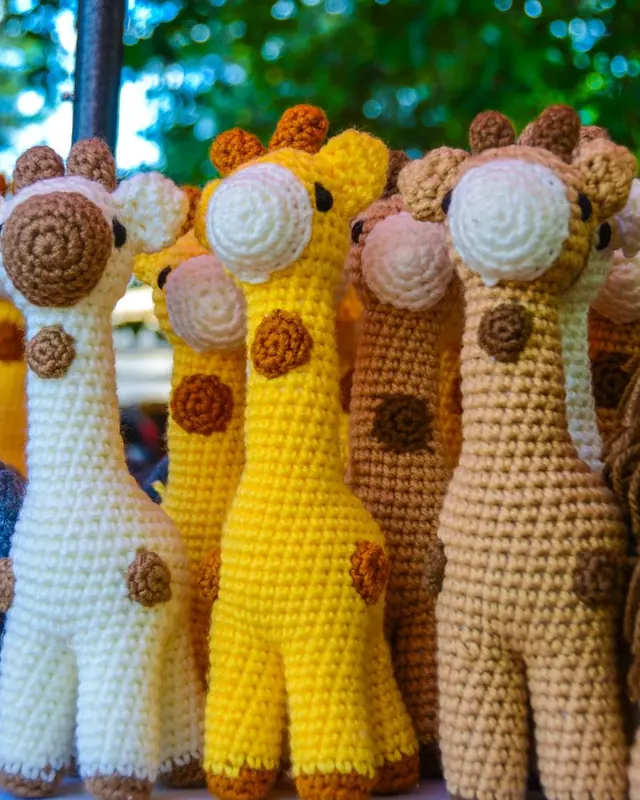 Three crocheted giraffes in cream, yellow and tan standing in front of green trees