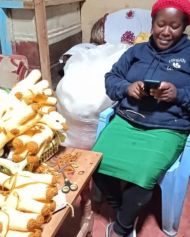 Woman in a red hat and navy hoodie looking at her phone on a plastic chair beside a table of crocheted giraffe pieces and scissors