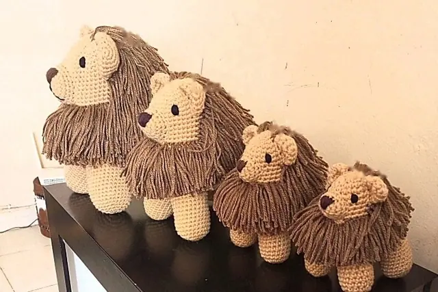Four crocheted tan lions with brown manes in a row on a black table, from largest at the left to smallest at the right, against a cream wall