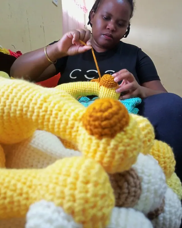 Woman in a black t-shirt crocheting with a hook, with yellow and cream crocheted giraffe pieces in the foreground