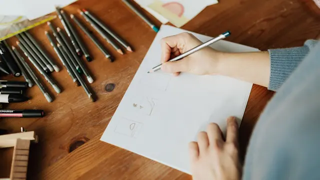 A hand sketching on paper beside coloured pencils on a wooden table