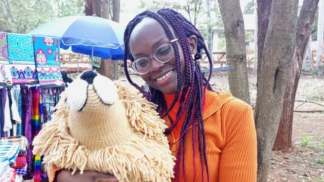 Smiling woman with purple braids and an orange top holding a large crocheted lion at an outdoor market with kitenge cloths behind her
