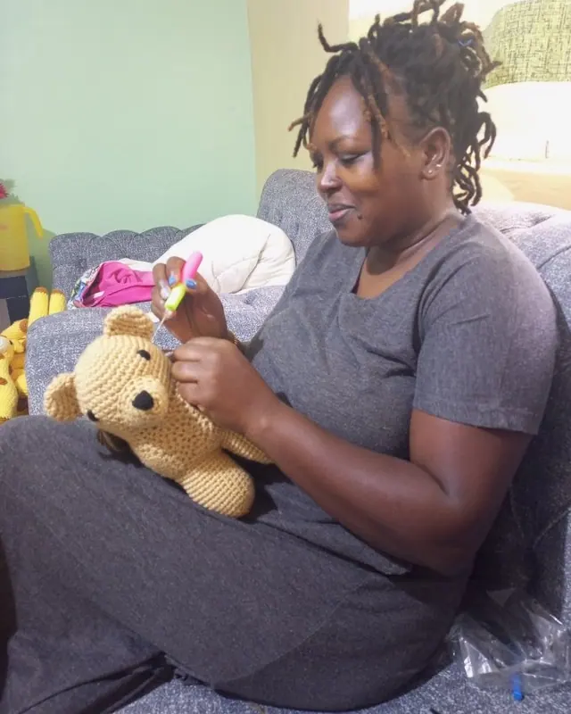 Woman with locs on a grey sofa sewing the face of a tan crocheted bear on her lap
