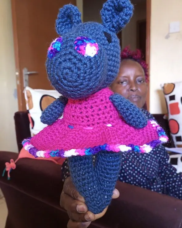 Hand holding up a navy crocheted hippo wearing a magenta dress, with a seated woman behind