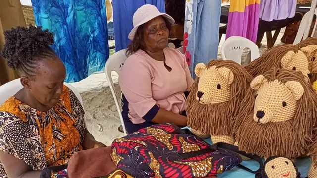 Two women at a stall on sand with crocheted lions and dolls on a blue table