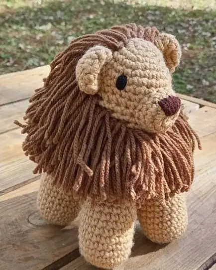 Crocheted lion with a tan body and a long brown yarn mane standing on a wooden pallet in the sun