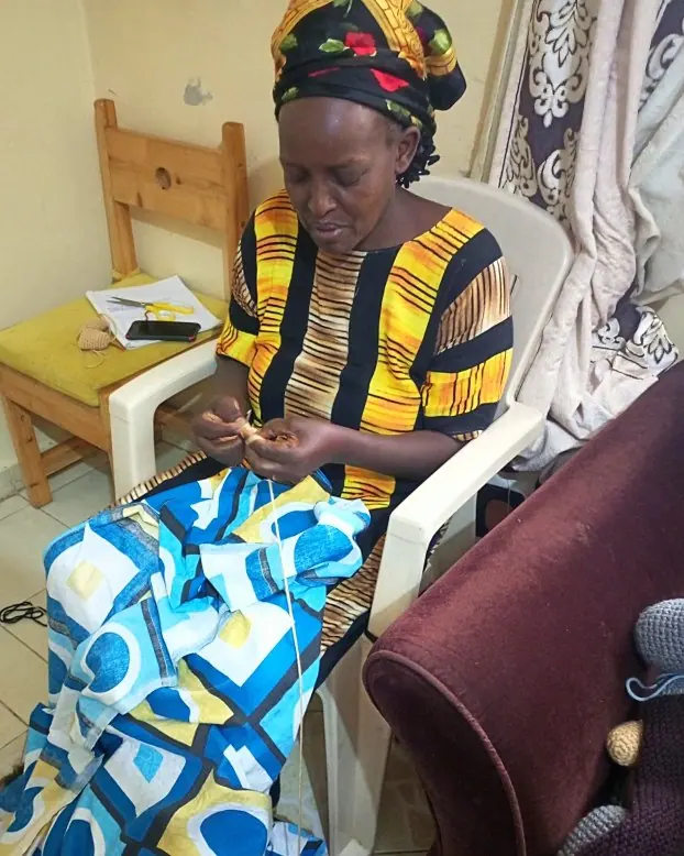 Woman in a patterned yellow and black top and kitenge skirt crocheting on a chair, with a crocheted animal waiting beside her