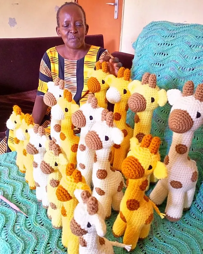 Woman in a striped yellow dress seated behind about fifteen crocheted yellow and cream giraffes on a teal blanket