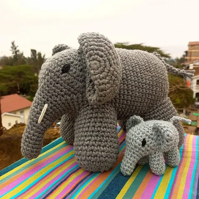 A large dark grey crocheted elephant with a white tusk and a small light grey baby elephant sitting on a striped cloth with town rooftops behind.
