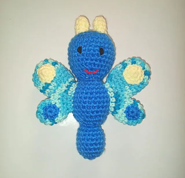 A blue crocheted butterfly with turquoise patterned wings, yellow antennae and a red smile, laid flat on a pale background.