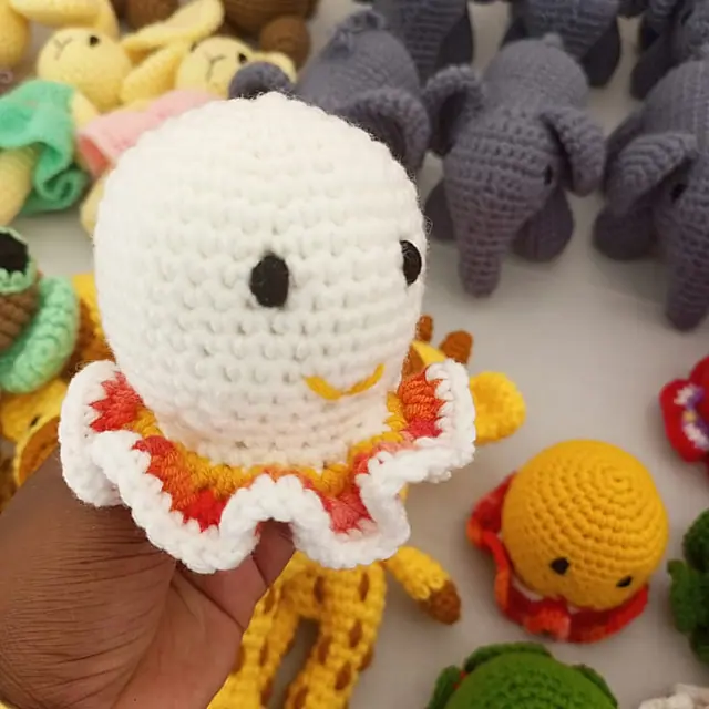 A hand holding a white crocheted toy with an orange and red ruffled edge, surrounded by grey elephants, yellow giraffes and green animals.