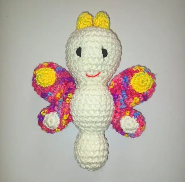 A cream crocheted butterfly with pink, purple and blue speckled wings and yellow antennae on a plain background.