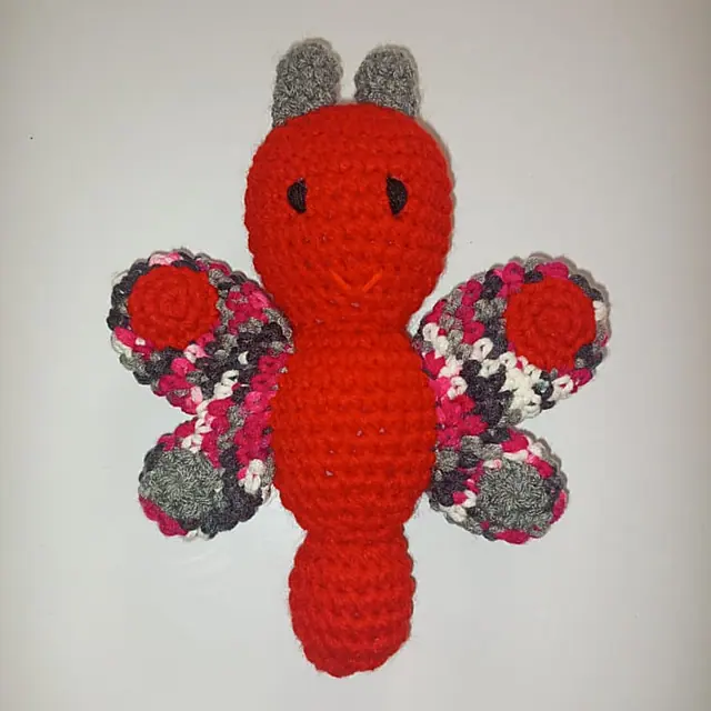Red crocheted butterfly toy with grey antennae and pink, black and white speckled wings, laid flat on a plain light background.