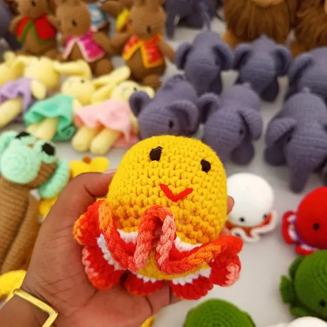 A hand holding a small yellow crocheted toy with a red and orange ruffled skirt, in front of rows of grey elephants, brown rabbits and other crocheted animals.