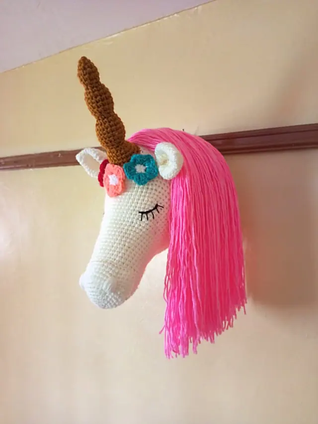 Crocheted unicorn head mounted on a yellow wall, with a cream face, brown horn, small flowers and a long bright pink yarn mane.