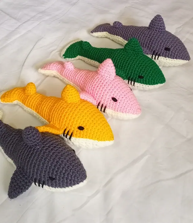 Five crocheted sharks in yellow, pink, green, slate purple and charcoal grey with cream bellies laid in a row on a white sheet.