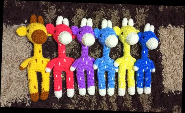 Six slim crocheted giraffes in yellow with brown spots, red, purple, light blue, pale yellow and royal blue lined up on a shaggy rug.