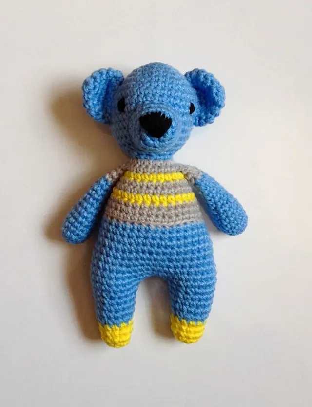 Sky blue crocheted bear with round ears, black nose, grey and yellow striped top and yellow feet, lying flat on a white background.