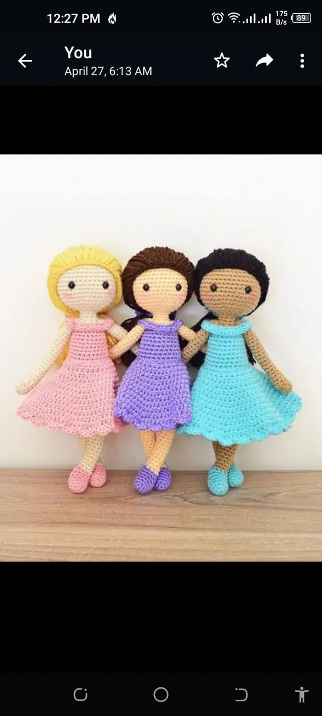 Three crocheted dolls in pink, purple and aqua dresses standing side by side on a wooden shelf.