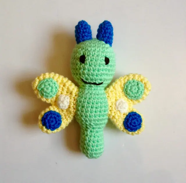 Crocheted butterfly with a mint green body, blue antennae and pale yellow wings with green, white and blue spots on a plain background.