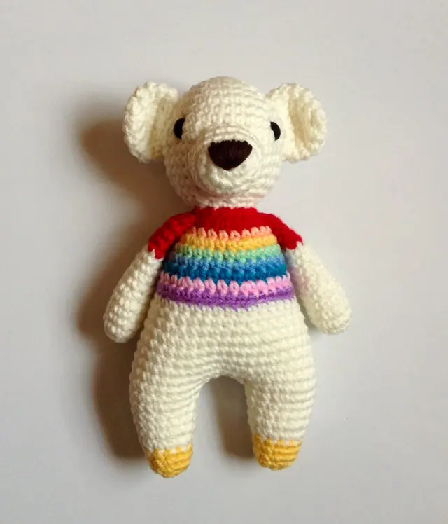 Cream crocheted bear with a rainbow striped top, red shoulders and yellow feet, on a plain grey background