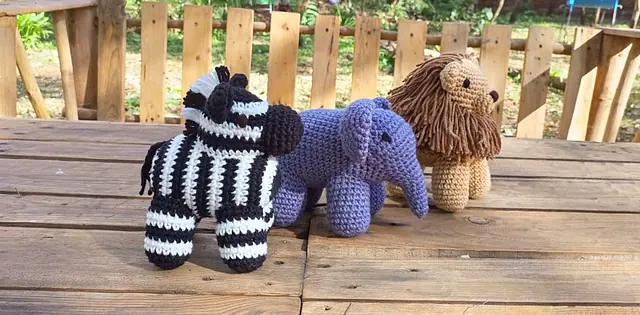 Crocheted black and white zebra, lavender elephant and tan lion with a brown yarn mane on a wooden table outdoors
