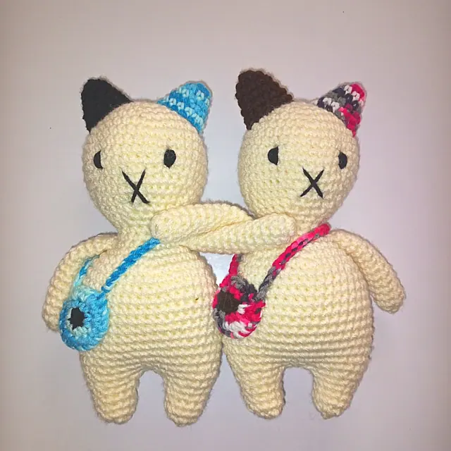 Two cream crocheted cats with linked arms, one with a turquoise bag and one with a pink variegated bag