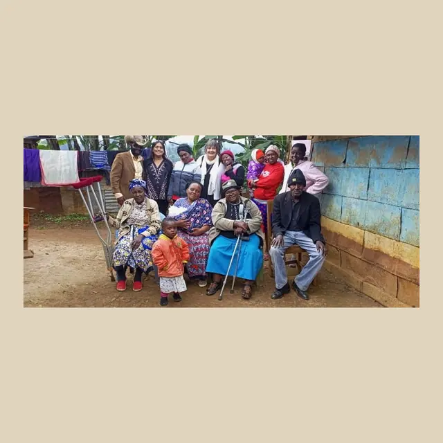 Group of about thirteen adults and children posing outdoors in front of a blue wall, some seated, including elderly people with walking sticks and a toddler in an orange jacket