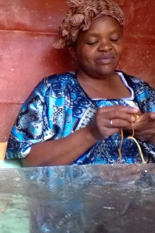 Woman in a blue patterned top and a brown head wrap crocheting with orange yarn, her eyes looking down