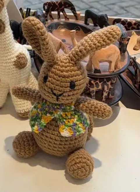 A brown crocheted rabbit with long ears and a multicoloured crocheted collar sits on a table in front of carved wooden safari animals.