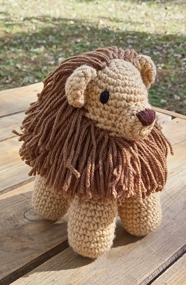 A tan crocheted lion with a long brown yarn mane and a dark maroon nose stands on a wooden table outdoors.
