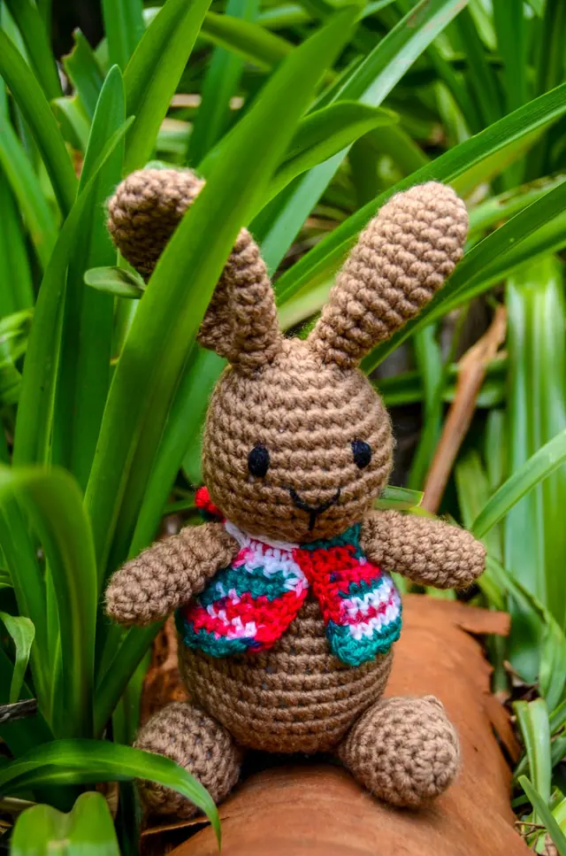 Brown crocheted rabbit with long ears and a red, teal and white striped vest, waving from a log among green leaves