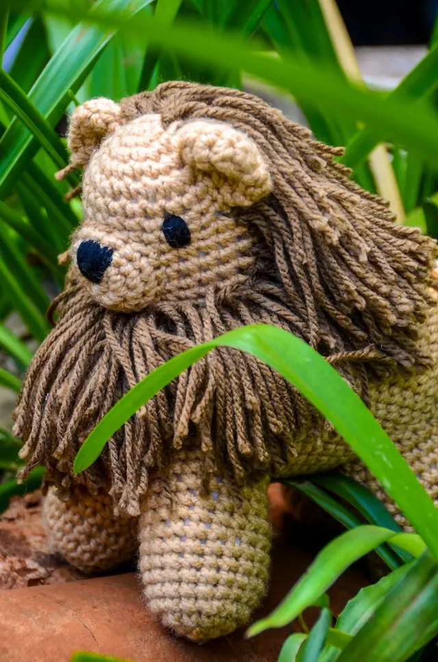 Tan crocheted lion with a long brown yarn mane standing among green leaves