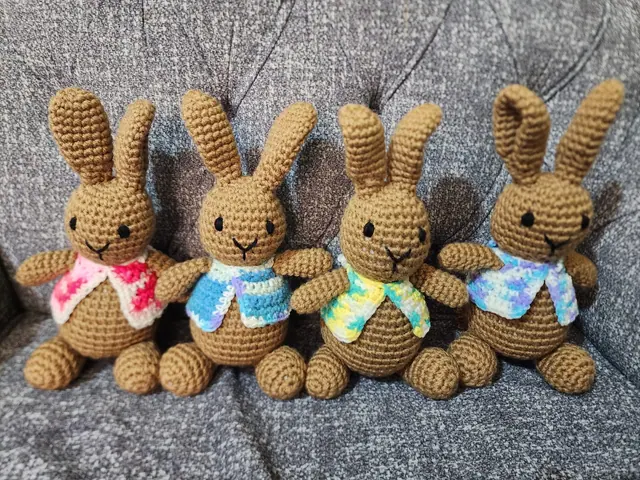 Four brown crocheted rabbits in pink, blue, yellow and lilac variegated vests sitting in a row on a grey sofa
