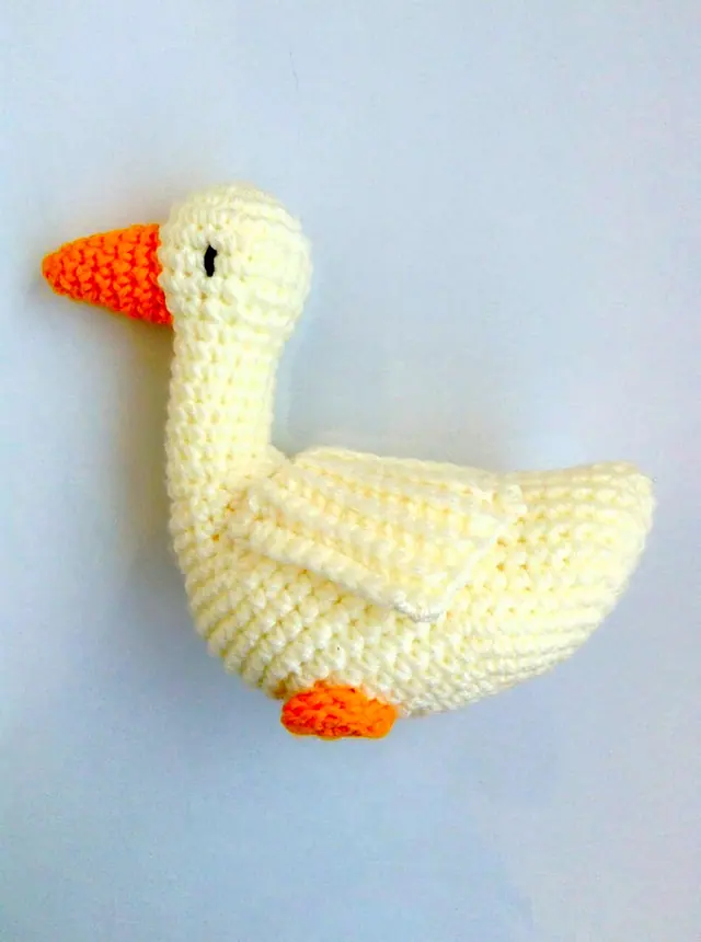 Cream coloured crocheted bird with an orange beak and foot on a plain pale background