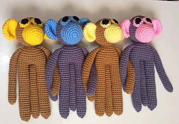 Four crocheted monkeys lying side by side, two brown with yellow ears and muzzles and two purple with blue and pink ears and muzzles.