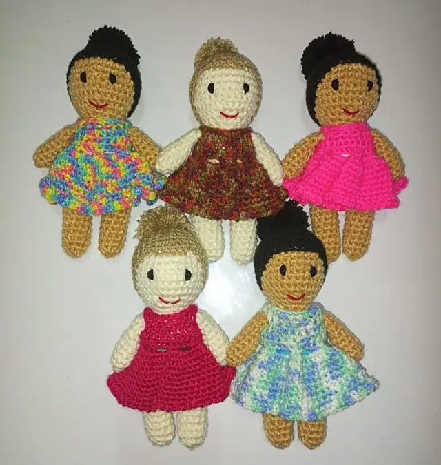 Five crocheted dolls in colourful crocheted dresses, three with tan skin and black hair buns and two with cream skin and tan hair buns.