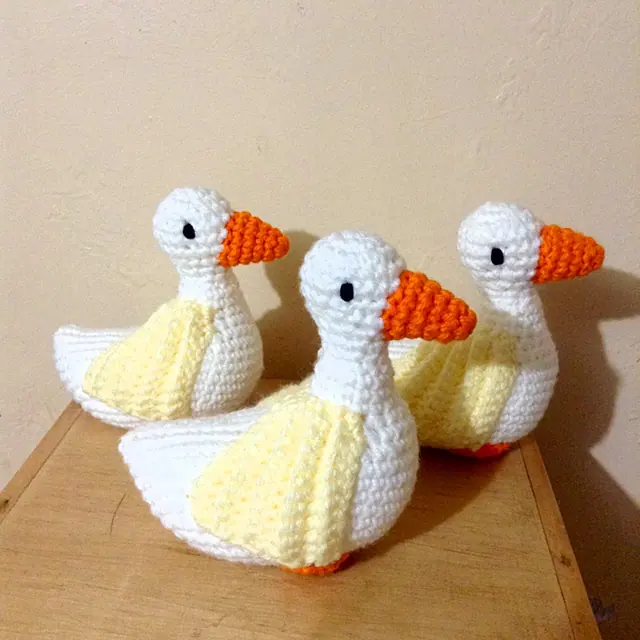 Three crocheted ducks with white bodies, pale yellow wings and orange beaks sitting on a wooden table.