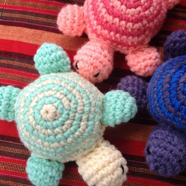Three crocheted turtles seen from above on a red striped cloth, one mint green and cream, one coral pink and one blue and purple.