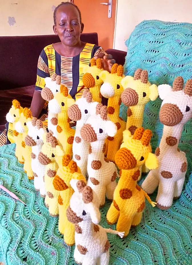 Woman in a striped yellow dress seated behind about fifteen crocheted yellow and cream giraffes on a teal blanket
