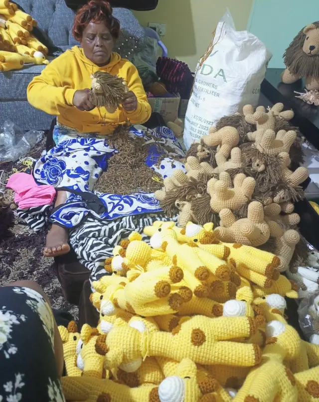 Older woman in a yellow hoodie hand finishing a lion mane surrounded by piles of crocheted giraffes and lion bodies