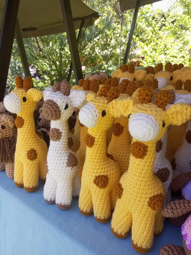 Rows of yellow and cream crocheted giraffes on a pale blue cloth under a canopy with green trees behind