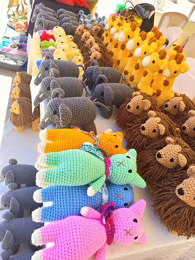 Market table with rows of crocheted giraffes, grey elephants and lions, and four long cats in orange, mint, blue and pink lying at the front
