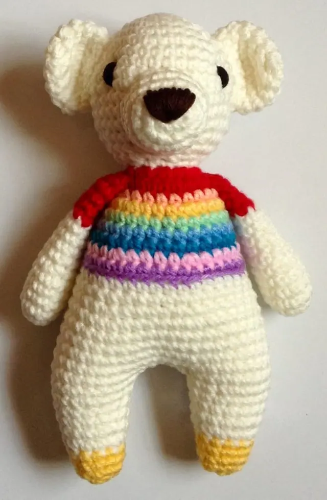Cream crocheted bear with a brown nose, a red-shouldered rainbow striped top and yellow feet, on a plain pale background.