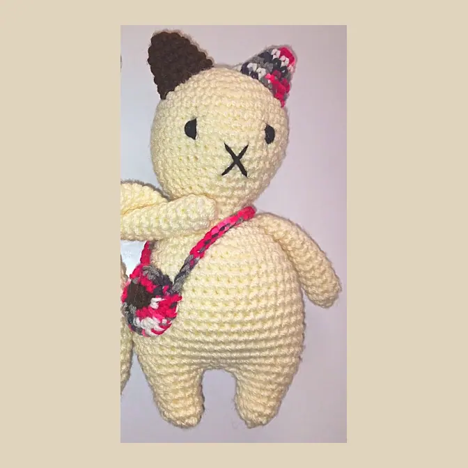 Cream crocheted cat with a red and grey speckled ear tip, brown ear and a small red and grey shoulder bag, with its friend close beside it