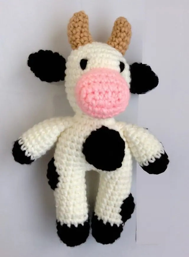 Black and white crocheted cow standing upright, with a cream body, black patches, black ears, tan horns and a large pink nose, on a pale grey background.