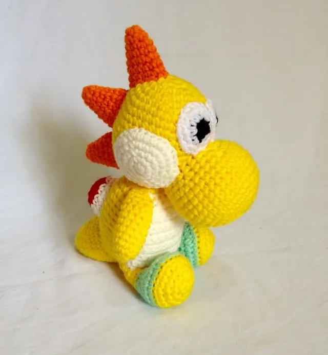 Sitting yellow crocheted dinosaur with orange spikes, large white eyes, cream cheek and belly and mint green feet, on a plain light background